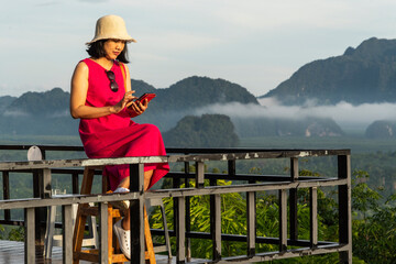  Asian woman wearing a pink dress sits neatly looking at the misty mountain view holding a smartphone on social networks and is happy to travel on vacation. 
