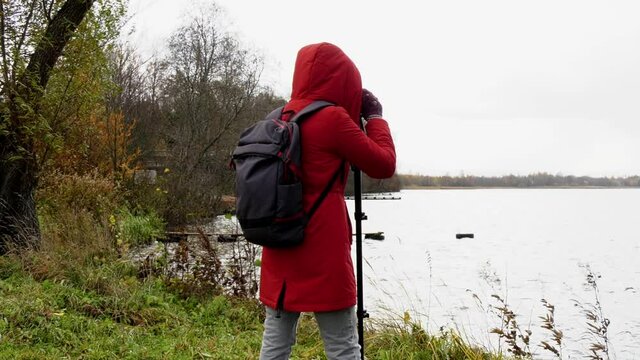 A woman with a backpack in a red jacket stands on the shore of the lake and takes pictures of the landscape.