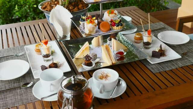 Afternoon tea in restaurant. Traditional English afternoon tea stand with selection of sweets - cakes, snacks, sandwiches and macaroons. Tea set and coffee with assortment of pastries in outdoor cafe