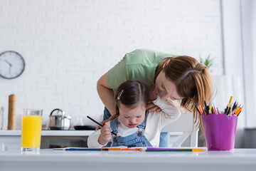 Smiling woman hugging daughter with down syndrome holding color pencil in kitchen.