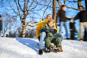 Young joyful father and his cheerful smiling little afro american or latin son in warm waterproof overalls slide down the crowdy snowy mountain on wooden sled on sunny winter christmas day. Holidays