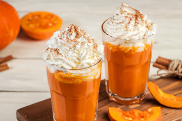 Spicy latte with pumpkin and whipped cream on wooden background. Hot coffee in glass mug and autumn leaves.