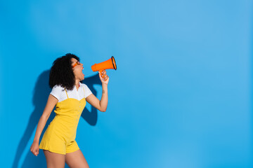 african american woman in yellow jumpsuit shouting in megaphone on blue