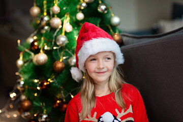 Cute blonde caucasian girl 7 years old sitting on floor near Christmas tree in Santa hat and smiling. Bedroom decorated with garlands. Xmas celebrate, happy new year 2022. Time to make wishes