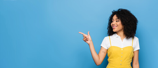 pleased african american woman looking away while pointing with finger on blue, banner