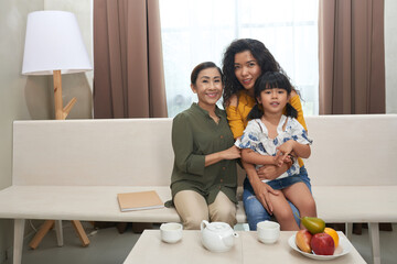 Portrait of smiling young Asian woman with daughter on knees sitting on sofa with senior mother in...