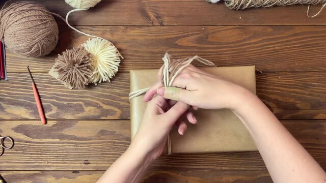 Women's hands ties gift box in craft paper with twine. Yarn and pom-poms on wooden table. Top view of festive composition. Christmas decorations.