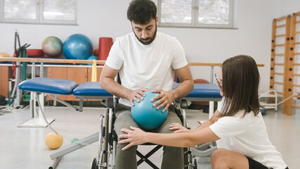 Man with disability in a wheelchair, doing an exercise with a smaller pilates ball at the physiotherapeutic rehabilitation center