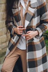 Closeup portrait of young beautiful fashionable woman wearing checkered long coat, beige pants and white blouse . Lady posing on city street.