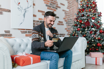 Playful man with a beard smiling sitting on the couch shopping online, buying Christmas presents,...