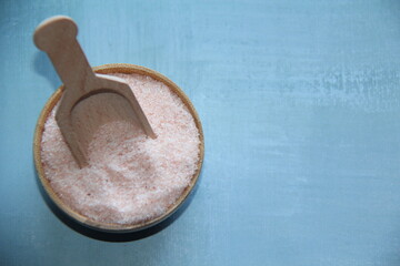 Pink Himalayan salt fine, ground or refined in a wooden bowl, with a wooden measuring spoon, isolated on a light blue background, top view.