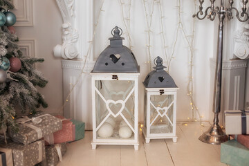 Christmas lanterns in decorated living room. Vintage decorative white lanterns in home. New year winter home decor. Large wooden lanterns with glowing garlands and gifts stand on floor in room. 
