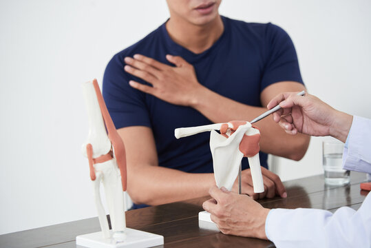 Horizontal shot of traumatologist sitting at desk in front of young Asian man consulting him about pain in shoulder joint