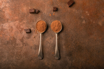 Chocolate-colored cocoa powder in two deep vintage spoons, broken pieces of chocolate around, on a textured rusty background.