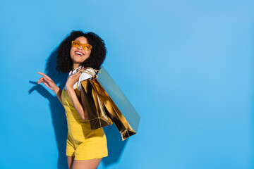 excited african american woman in yellow eyeglasses posing with shiny shopping bags on blue