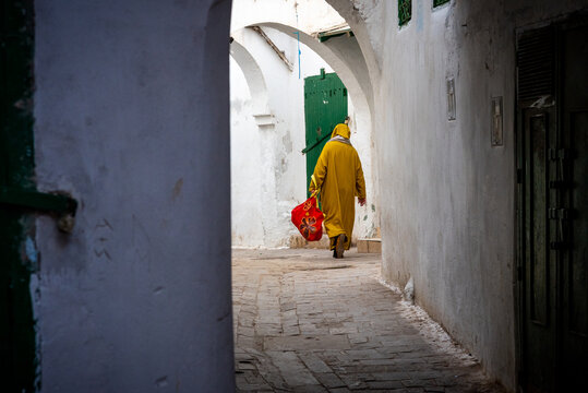 In the medina of Chefchaouen in Morocco. A man wearing the traditional Berber coat walks down the narrow street carrying a red bag