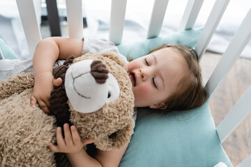 High angle view of child with down syndrome holding soft toy in baby crib. - obrazy, fototapety, plakaty