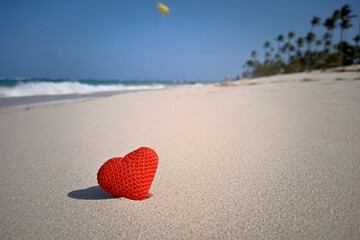 Red knitted heart in sand of tropical beach on background of coconut palm trees. Sea holidays, romantic love on paradise nature