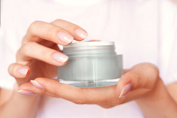 Female hands with natural pink manicure holding a transparent cream jar. Woman in white holds beauty product. Concept of body care and skin protection. Moisturizing lotion closeup. Facial skincare