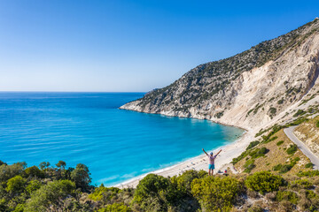 Fototapeta na wymiar Man tourist standing on top of a rock, raising hands with an exciting feeling of freedom, looking at Myrtos Beach. Cephalonia island, Greece