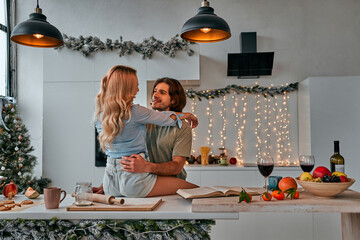Kitchen love stories. A gorgeous woman is hugging her happy husband while sitting on a kitchen countertops. Preparation for the Christmas holidays.
