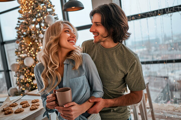 Couple casual stylish lovers are drinking coffee in the kitchen in new year morning. Young woman and her husband with a cups in them hands are smiling happy. Lifestyle christmas concept.