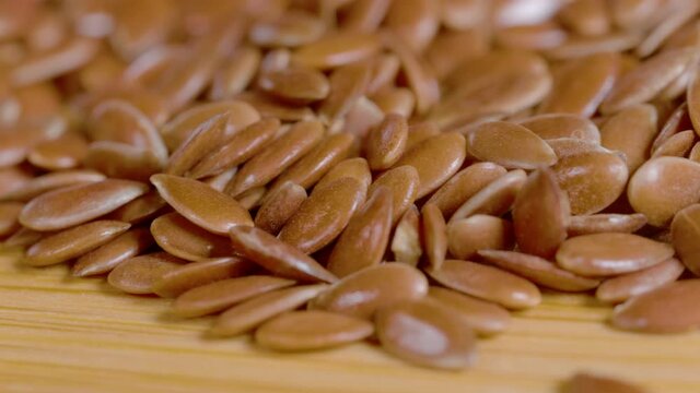 MACRO, DOF: High definition shot of a small pile of reddish brown flaxseeds. A heap of linseeds lies scattered across the wooden dining table surface. Shiny linseeds lie on the wooden countertop.