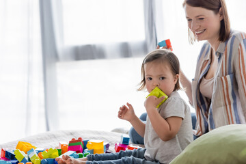 Kid with down syndrome playing building blocks near smiling mother on bed.
