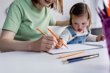 Blurred kid with down syndrome drawing on paper near mom in kitchen.