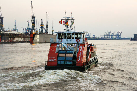 Passenger ferry sightseeing tour boat leaving from St. Pauli's Piers or Landungsbrücken with Blohm and Voss shipyards and sunset sky in background at port of Hamburg. 