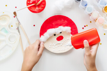 The child glues the parts Santa made from paper plate. Handmade. Project of children's creativity, handicrafts, crafts for kids. Preparation for christmas. Decoration.