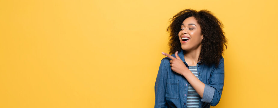 laughing african american woman looking away and pointing with finger isolated on yellow, banner