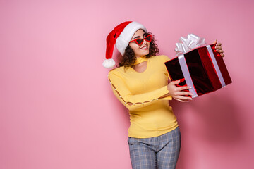 Smiling curly girl in Santa's hat, sunglasses holds giftbox while stands on pink background New Year