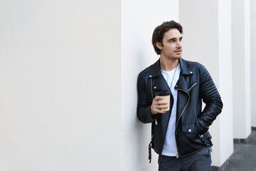 Stylish man in leather jacket holding coffee to go near while wall 