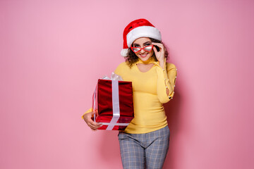 Happy curly girl in Santa's hat, glasses holds present while stands on pink background. New Year
