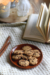 Fototapeta na wymiar Plate of chocolate chip cookies, cinnamon sticks, soft blanket, open book and lit candles. Hygge at home. Selective focus.