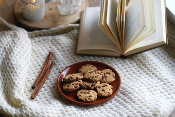 Plate of chocolate chip cookies, cinnamon sticks, soft blanket, open book and lit candles. Hygge at home. Selective focus.