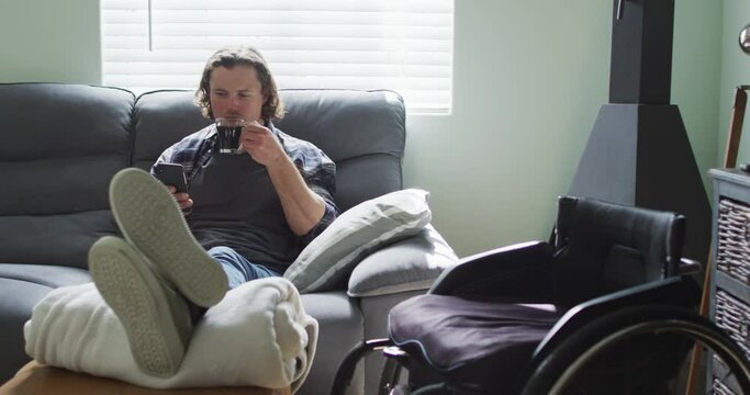 Happy caucasian disabled man sitting on sofa in living room using smartphone and drinking coffee