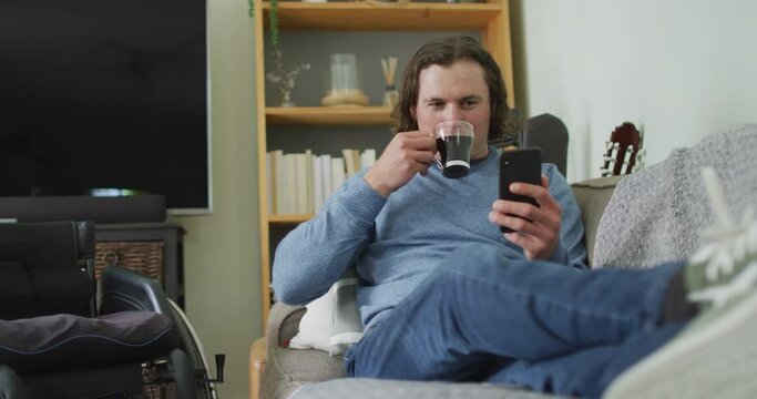 Happy caucasian disabled man sitting on sofa in living room using smartphone and drinking coffee