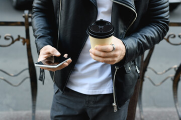 Cropped view of man in leather jacket using smartphone and holding coffee on urban street 