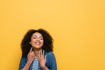 happy african american woman in denim shirt holding hands near neck while posing isolated on yellow