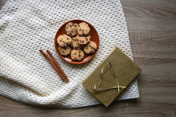 Plate of chocolate chip cookies, knitted blanket, book, reading glasses and cinnamon sticks. Hygge at home. Flat lay.