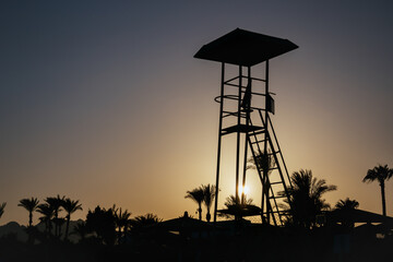 Beautiful sunset sky with silhouette lifeguard tower and coconut palm trees. Abstract nature and travel background. Egypt summer. Observation tower in the evening