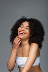 excited african american woman in white top laughing with closed eyes isolated on grey