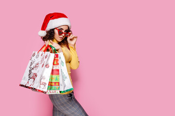 Curly girl in Santa's hat and sunglasses holds shopping bags while stands on pink background. Sales