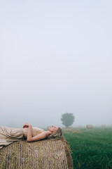 Naklejka na ściany i meble Young woman in beige dress lying on haystacks at twilight time