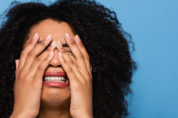 upset african american woman obscuring face with hands while crying isolated on blue