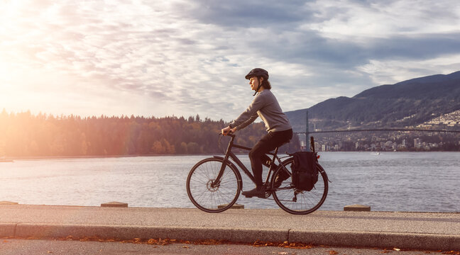 White Caucasian Adult Woman riding a bicycle on Seawall in Stanley Park. Sunset Sky Art Render. Downtown Vancouver, British Columbia, Canada.