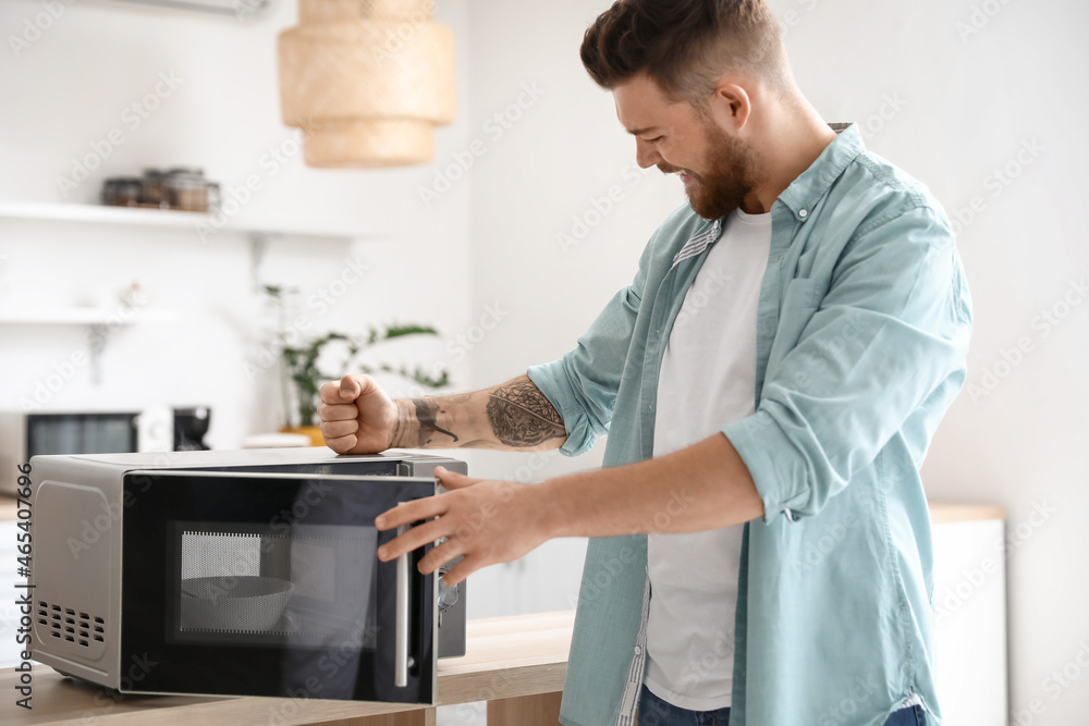 Wall mural Angry young man near microwave oven in kitchen