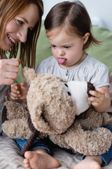 Smiling woman playing with baby daughter with down syndrome and soft toy on bed.